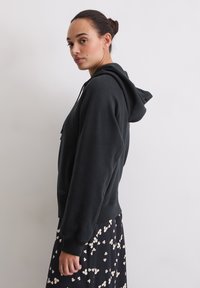 Felpa nera con cappuccio e tasca a marsupio, dal taglio rilassato. Indossata sopra una gonna nera a fantasia floreale.