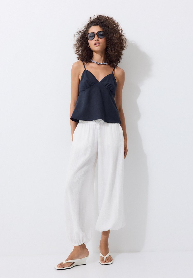Femme aux cheveux bouclés portant des lunettes de soleil foncées, un top noir sans manches, un pantalon blanc ample et des sandales blanches, debout contre un mur blanc.