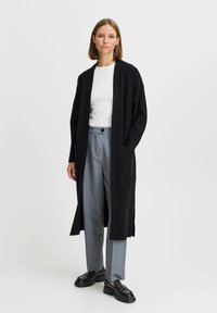 Svart lång cardigan med avslappnad passform, två stora framfickor, i kombination med grå byxor och svarta kraftiga skor. Slät textur.