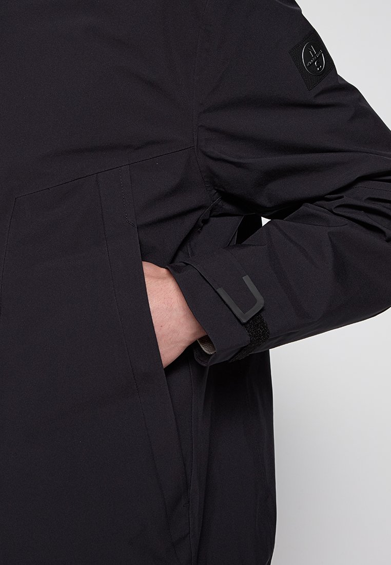 Giacca nera impermeabile con finitura opaca, dotata di tasca con zip, polsini in velcro e una patch con logo sulla manica.