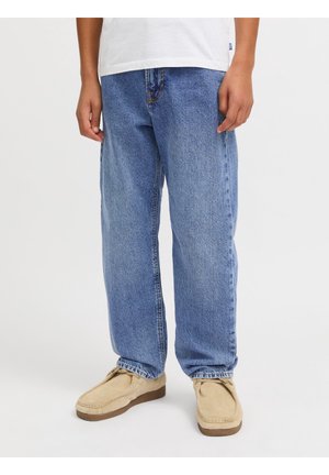 Personne portant un jean denim bleu ample, des mocassins beiges et une chemise blanche partiellement visible à la taille.