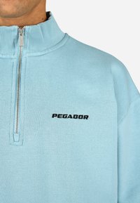 Gaiši zils ceturtdaļas rāvējsvētrojums, izgatavots no mīkstas auduma, ar ribotu apkakli un melnu "PEGADOR" logotipu uz krūtīm.