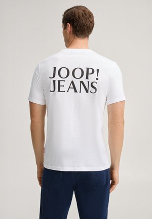 Homme de dos, portant un T-shirt blanc avec "JOOP! JEANS" en grandes lettres noires dans le dos et un jean bleu foncé dans un cadre neutre.