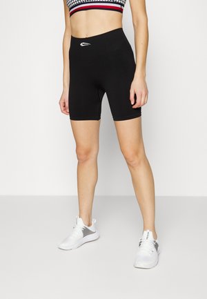 Kvinde iført sorte biker-shorts med høj talje, mønstret crop top og hvide træningssko, stående mod en ensfarvet baggrund.