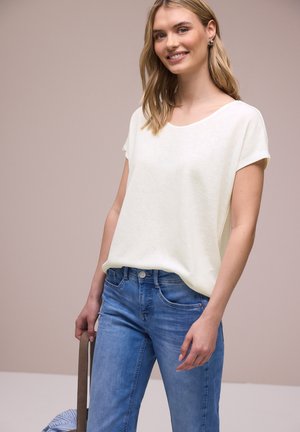 Smilende kvinde iført en hvid kortærmet top og blå jeans, der holder en stribet taske ved sin side mod en neutral baggrund.