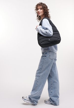 Donna con capelli ricci, felpa azzurra e jeans larghi, che porta una grande borsa a tracolla nera imbottita, indossando sneakers bianche e grigie.