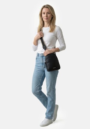 Blonde Frau, die ein weißes Langarmshirt, hellblaue Jeans, weiße Sneakers trägt und eine schwarze Umhängetasche mit breitem Riemen bei sich hat.