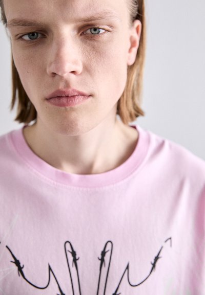 T-shirt en coton rose clair avec un design graphique comportant des lignes et des détails noirs sur le devant. Encolure ras du cou et texture douce.