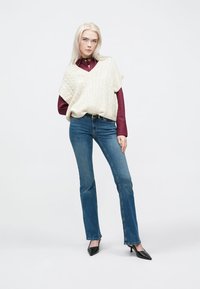 Svetla pletena brezrokavnik preko burgundske srajce, v kombinaciji z modrimi kavbojkami in črnimi čevlji s koničastim prstom. Gladka tekstura in sproščen kroj.