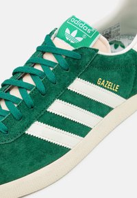 Green suede sneaker with white three-stripe design, gold "GAZELLE" lettering, and green tongue label featuring the Adidas logo.