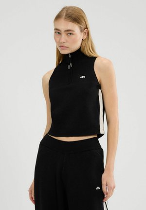 Jonge vrouw die een mouwloze zwarte zip-up crop top en bijpassende zwarte broek met witte zijstrepen draagt, staand tegen een effen achtergrond.