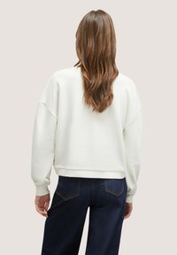 Sudadera blanca texturizada con puños y dobladillo acanalados, mostrada desde la espalda, combinada con unos vaqueros de azul oscuro.
