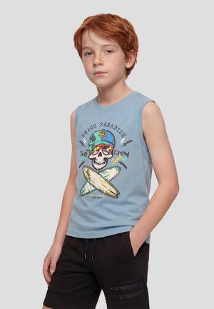 Niño pelirrojo con camiseta sin mangas azul claro con diseño de calavera con gafas de sol y tablas de surf, pantalones cortos negros, manos en los bolsillos.
