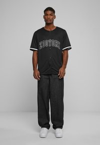 Chemise noire à manches courtes et boutonnée avec le logo "ZOORYORK" en blanc, associée à un pantalon ample gris foncé et des baskets blanches.