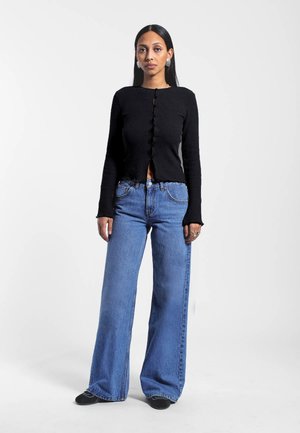 Femme debout portant un haut noir côtelé à manches longues avec des boutons au centre et un jean bleu à jambes larges avec des chaussures noires sur fond blanc.