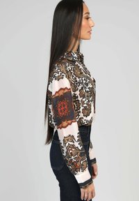 Blouse à manches longues en tissu de soie présentant un motif orné multicolore aux teintes de marron, orange et crème. Col classique et fermeture à boutons.