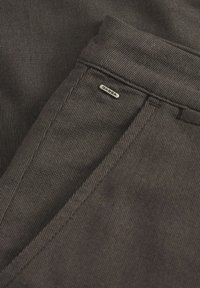 Donkergrijs gestreepte stof toont een close-up van een broek met een Gabba-logo etikett. Gestructureerd materiaal met zichtbare stiksels en details.