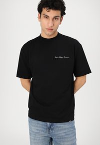 Black cotton t-shirt with a relaxed fit, featuring short sleeves and white embroidered text on the left chest. Pairing with light blue jeans.