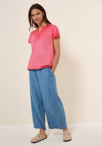 Crop top rosso a maniche corte con scollatura a V e vestibilità rilassata, abbinato a pantaloni larghi azzurri e scarpe slip-on beige.