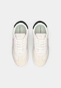 O pereche de adidași Calvin Klein bej deschis, cu șireturi albe și accente negre la călcâi, văzuți dintr-o parte de sus pe un fundal simplu.