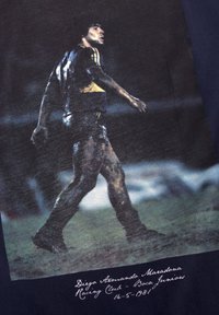 T-shirt met een grafisch ontwerp van een voetballer in een blauw-geel shirt, modderige shorts, met donker haar, die op een veld loopt, en tekst daaronder.