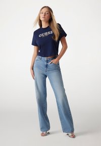 Marinblå crop top med "GUESS"-logga, matchad med ljusblå högmidjade vida jeans med slitdetaljer och silverfärgade sandaler.
