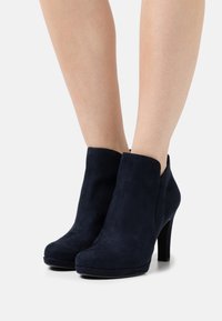 Botas de tobillo de ante azul marino con parte superior curva, puntera afilada y tacón de 10 cm. Textura suave y diseño minimalista; sin herrajes ni adornos visibles.