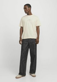Helles beiges Baumwoll-T-Shirt mit schwarzem "MATISSE"-Druck, kombiniert mit dunklen, lockeren Jeans aus Denim und beigen Sneakern mit strukturierten Akzenten.
