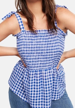 Kobieta ubrana w niebiesko-biały kraciasty top bez rękawów z wiązanymi na kokardę ramiączkami, stojąca z rękami na biodrach.