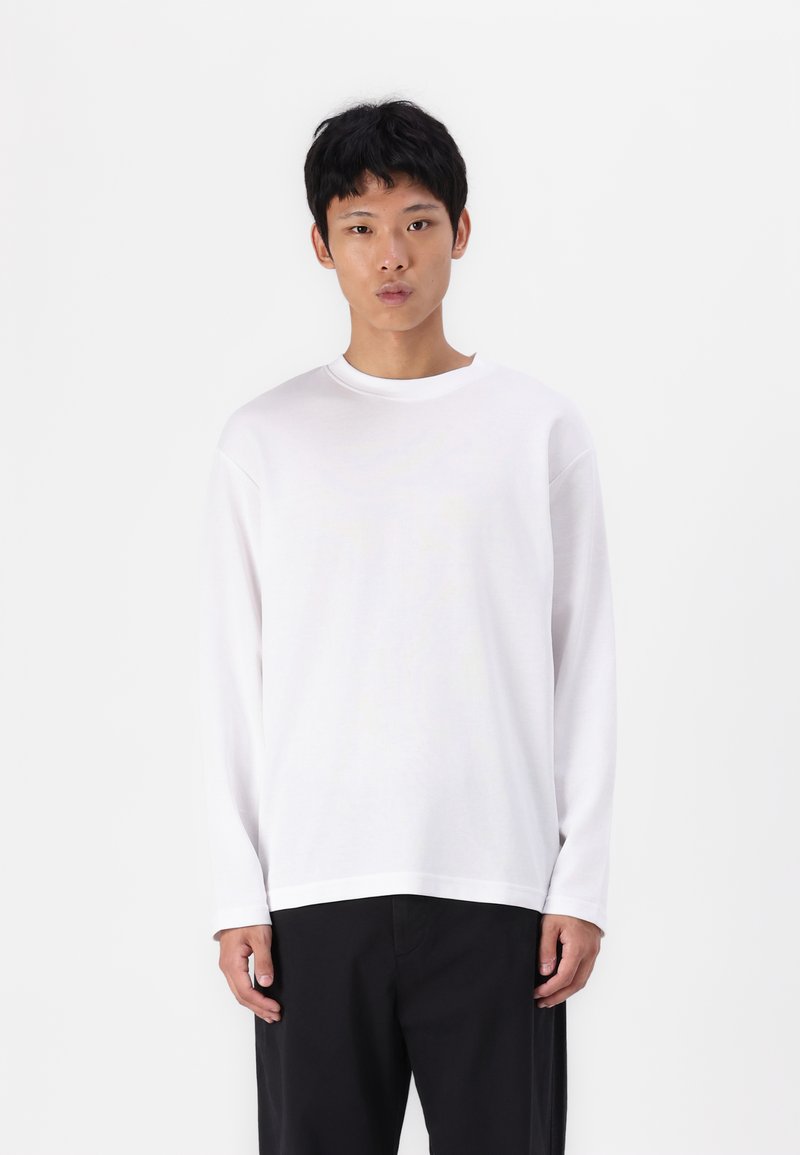 Λευκό μακρυμάνικο μπλουζάκι με στρογγυλή λαιμόκοψη και χαλαρούς ώμους, κατασκευασμένο από απαλό ύφασμα, συνδυασμένο με μαύρα παντελόνια. Απλός, μινιμαλιστικός σχεδιασμός.