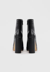 Pár černých kožených kotníkových bot na vysokém podpatku s blokovými podpatky a zadními zipy na bílém pozadí.