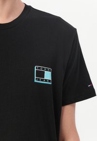 T-shirt noir en coton avec un logo carré turquoise et le texte "TOMMY JEANS" sur la poitrine gauche. Manches courtes avec un petit accent brodé.