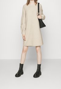 Beige pletené šaty svetrového typu s volným střihem, žebrovanou texturou a dlouhými rukávy, doplněné černou kabelkou a masivními černými kotníkovými botami.