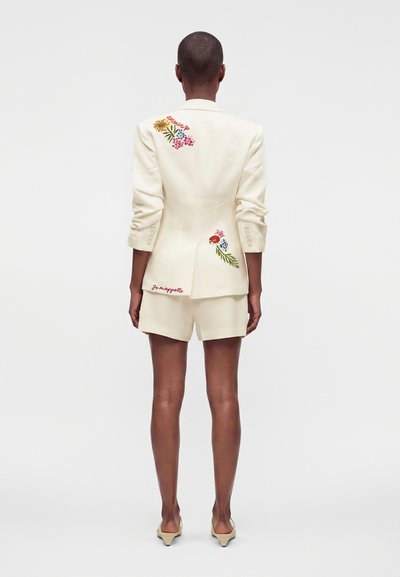 Personne debout, dos tourné vers la caméra, portant un blazer crème et un short ornés de broderies florales colorées et des mots « Je m’appelle. »