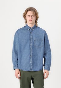 Cămașă din denim albastru deschis, cu guler cu nasturi, buzunar pe piept, mâneci lungi și croială lejeră. Textură netedă cu detalii de cusătură vizibile.