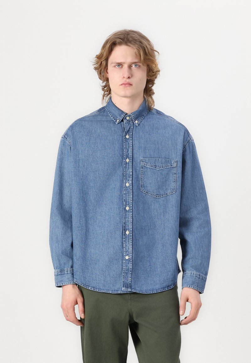 Cămașă din denim albastru deschis, cu guler cu nasturi, buzunar pe piept, mâneci lungi și croială lejeră. Textură netedă cu detalii de cusătură vizibile.