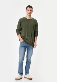 Løs pasform olivengrøn langærmet shirt med en kontrasterende halsudskæring, parret med lyseblå jeans og beige sneakers. Enkel design, blød tekstur.