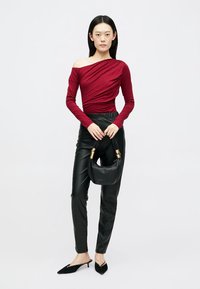 Blusa de manga longa bordeaux com design assimétrico, combinada com calças de imitação de couro pretas e uma pequena mala preta com detalhes em dourado.