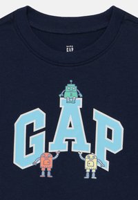 Μπλε βαμβακερό T-shirt με μεγάλο λογότυπο "GAP" σε ανοιχτό μπλε και τρεις καρτουνίστικους ρομπότ σε κόκκινο, πράσινο και κίτρινο, με τυπωμένες λεπτομέρειες.