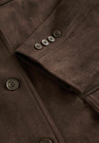 Veste en suedine marron avec un col à revers, fermeture à cinq boutons à l'avant et quatre boutons décoratifs sur la manche. Texture lisse.