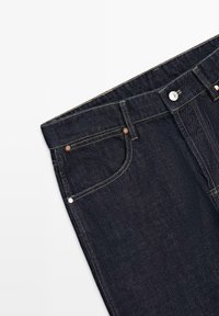 Dark blue denim jeans section showing front pockets, belt loops, copper rivets, and a metal button on the waistband against a white background.