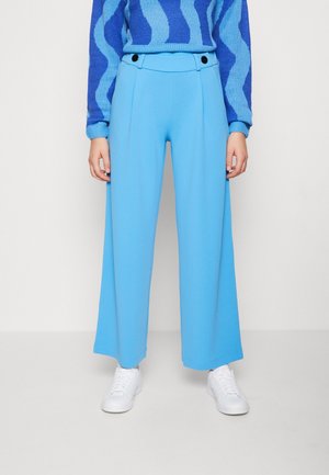 Person trägt weit geschnittene, hoch taillierte hellblaue Hose mit schwarzen Knöpfen und weißen Turnschuhen, kombiniert mit blau gemustertem Pullover.