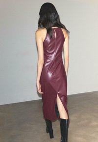 Un vestito in ecopelle bordeaux con silhouette aderente, caratterizzato da una fessura sul retro e un'apertura a chiave sul collo. Stivali neri alti completano il look.