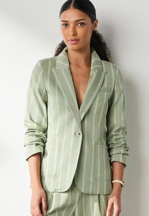 Woman with dark hair in a low ponytail wearing a light green pinstripe blazer with rolled sleeves and matching pants against a plain background.