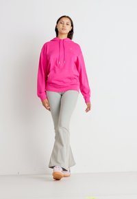 Felpa rosa con coulisse, tasca frontale, abbinata a pantaloni grigi chiari a campana e scarpe rosa con suola marrone. Sfondo neutro.