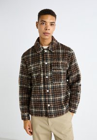 Braune karierte Jacke aus weichem Stoff mit kragen, vorderen Knöpfen und zwei Brusttaschen. Getragen über einem crèmefarbenen Pullover.
