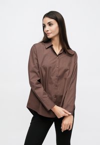 Chemise marron à boutons en coton, avec manches longues, col, boutons à l'avant et ourlet arrondi. Portée avec un pantalon noir.