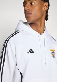 Felpa bianca con accenti neri, con tre strisce nere sulle maniche. Include il logo Adidas e il crest di Benfica con dettagli ricamati.