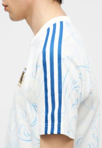 Osoba s bielou košeľou s svetlomodrými vzormi a tromi vertikálnymi modrými pruhmi na krátkom rukáve.