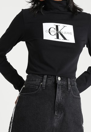 Schwarzer Langarm-Rollkragenpullover mit weißem Calvin Klein Jeans-Logo, kombiniert mit einem hoch taillierten dunkelblauen Jeansrock mit seitlichem Streifendetail.